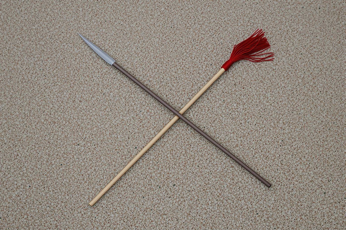 A steel-tipped spear and a wooden pole with a red tassel lying crossed on pale gravel